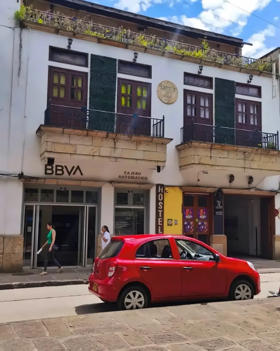 The building from the street: a white colonial facade with wooden balconies, the BBVA cash machine on the left and the vertical HOSTEL sign by a yellow pillar at the entrance on the right.