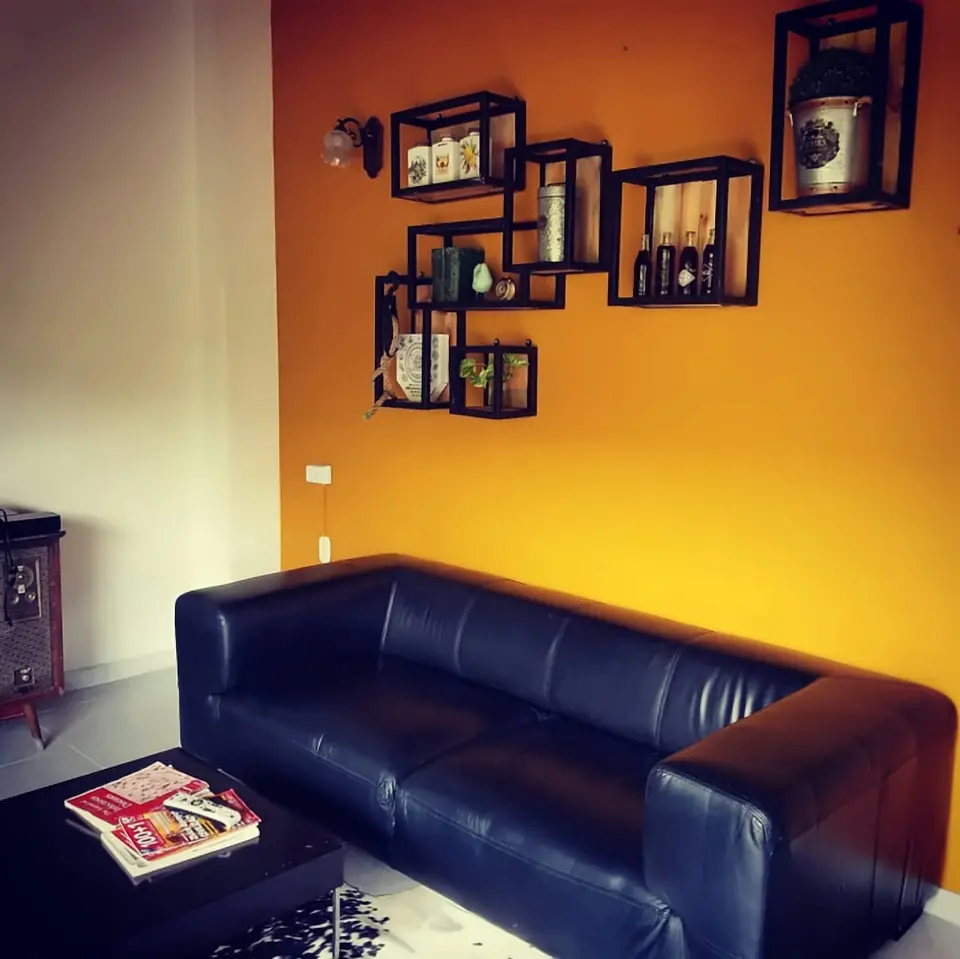 A black leather sofa against an ochre wall hung with black box shelves holding jars, plants and old bottles.