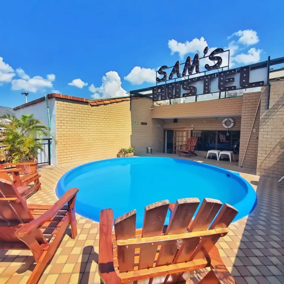 The rooftop by day: a small round blue pool on a tiled floor, wooden Adirondack chairs and the SAM’S HOSTEL letters against a blue sky.