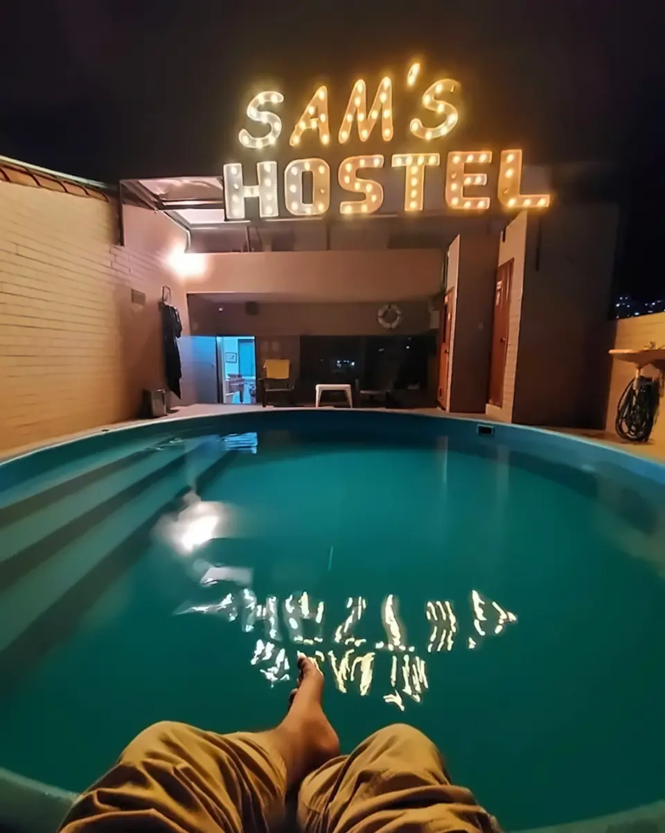 Night on the SamS rooftop: a guest’s feet at the edge of the small round pool, the lit SAM’S HOSTEL bulb sign reflected in turquoise water.
