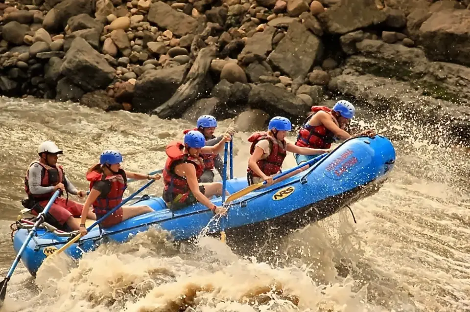 Rafters in blue helmets and red life jackets dig their paddles in as a blue raft punches through brown whitewater on the Río Suárez.