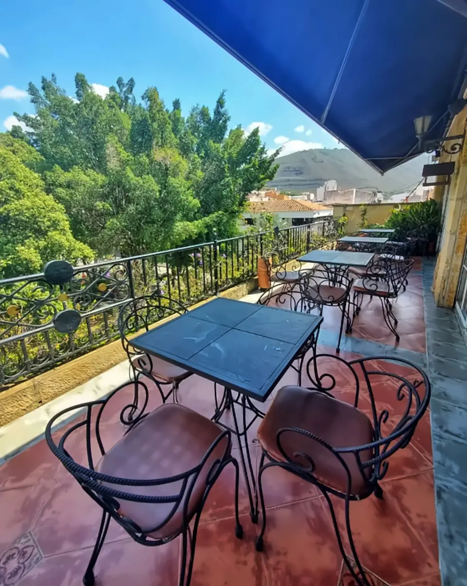 Terrace under a blue awning: iron chairs with leather seats around a slate-topped table, trees and a wooded hill beyond the railing.