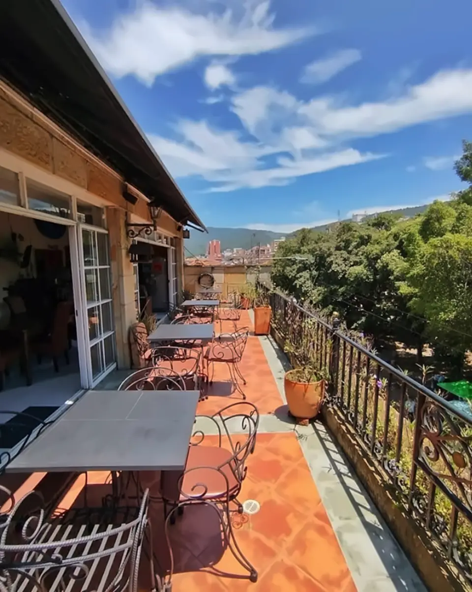 The sunny terrace: wrought-iron chairs and tables on orange tiles, potted plants along the railing and mountains beyond the rooftops.