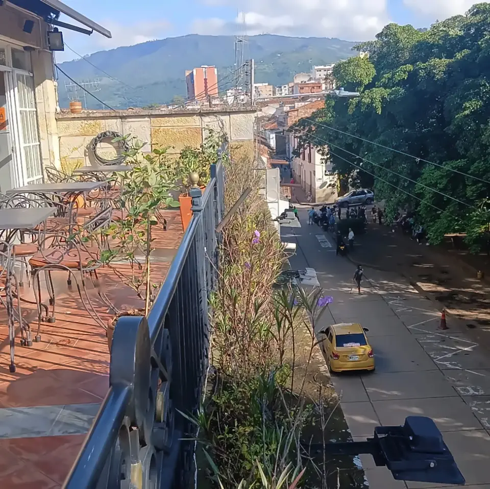 View from the hostel’s balcony: wrought-iron railing and plants, the street below with a yellow taxi, big trees on one side and green mountains behind the town.